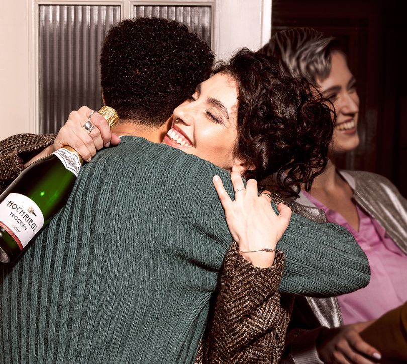Zwei Personen umarmen sich herzlich, wobei die eine Dame eine Flasche Hochriegl Trocken in der Hand hält