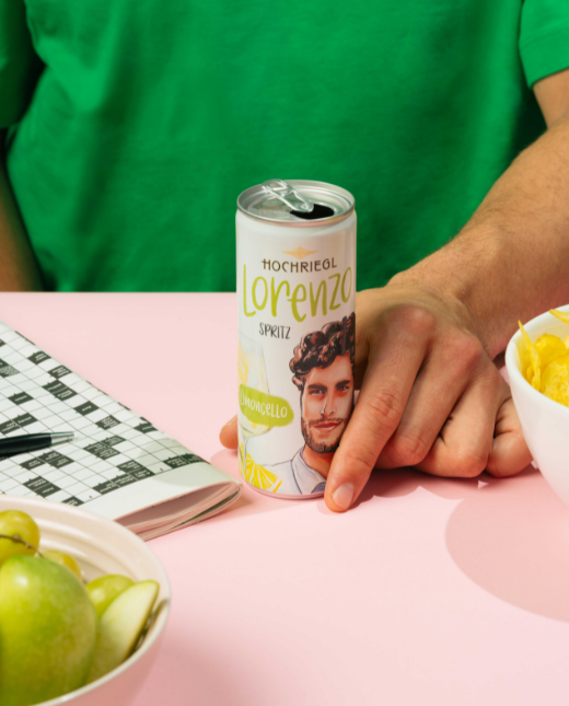 Man hält Lorenzo Dose in der Hand, am Tisch liegt ein Kreuzworträtsel, Äpfel und eine Schüssel mit Chips