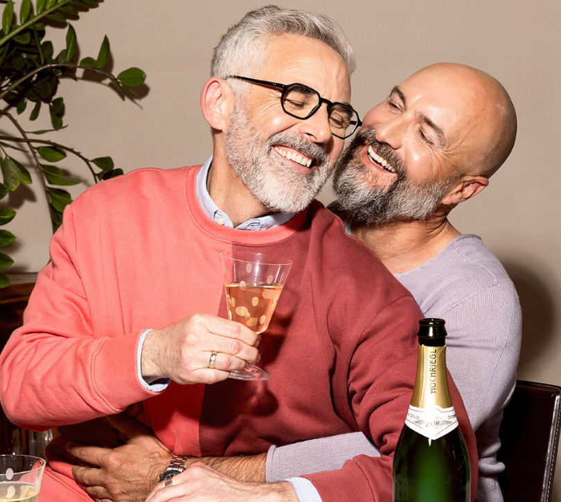 Ein Mann sitzt auf dem Schoß eines andern Mannes, sie lächeln sich an und trinken ein Glas Hochriegl