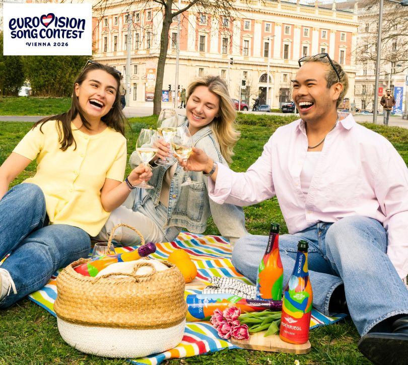 Drei junge Leute sitzen auf einer Picknickdecke im Gras und trinken ein Glas Sekt