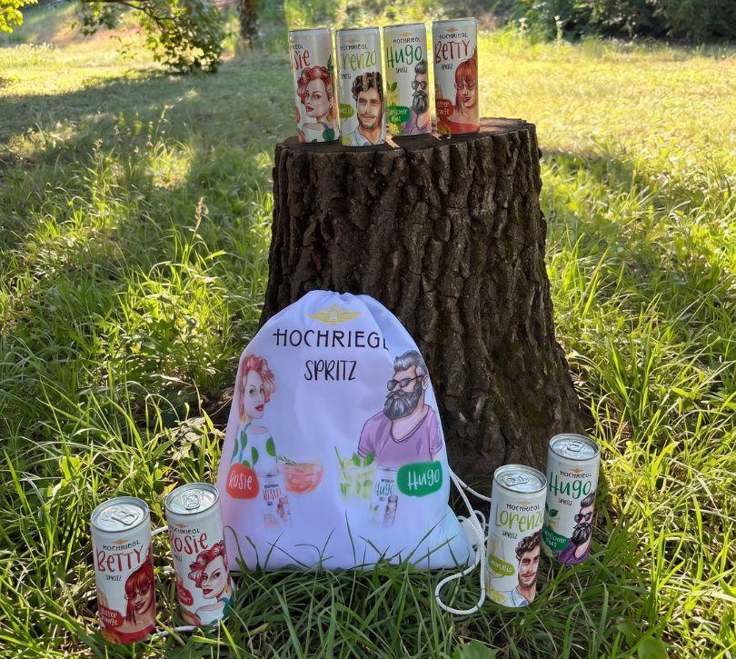 Spritz Dosen und Tasche liegen im Gras bzw. stehen am Baumstumpf