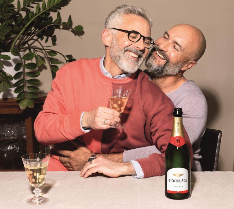 Ein Mann sitzt auf dem Schoß eines andern Mannes, sie lächeln sich an und trinken ein Glas Hochriegl