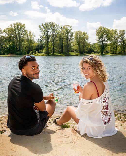 Ein junges Paar sitzt am Fluss und trinkt Hugo und Betty Dosen